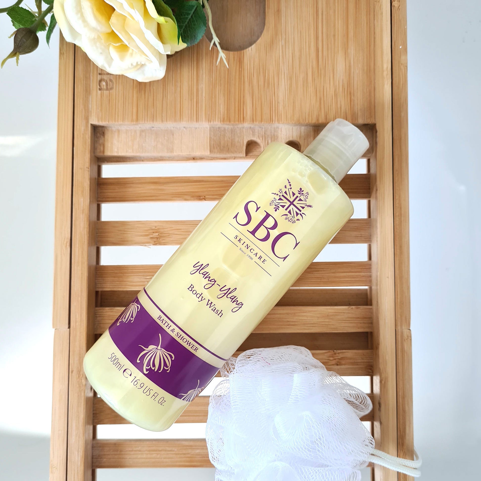 SBC Skincare's Ylang-Ylang Body Wash on a wooden bath caddy with flowers and a shower scrunchie
