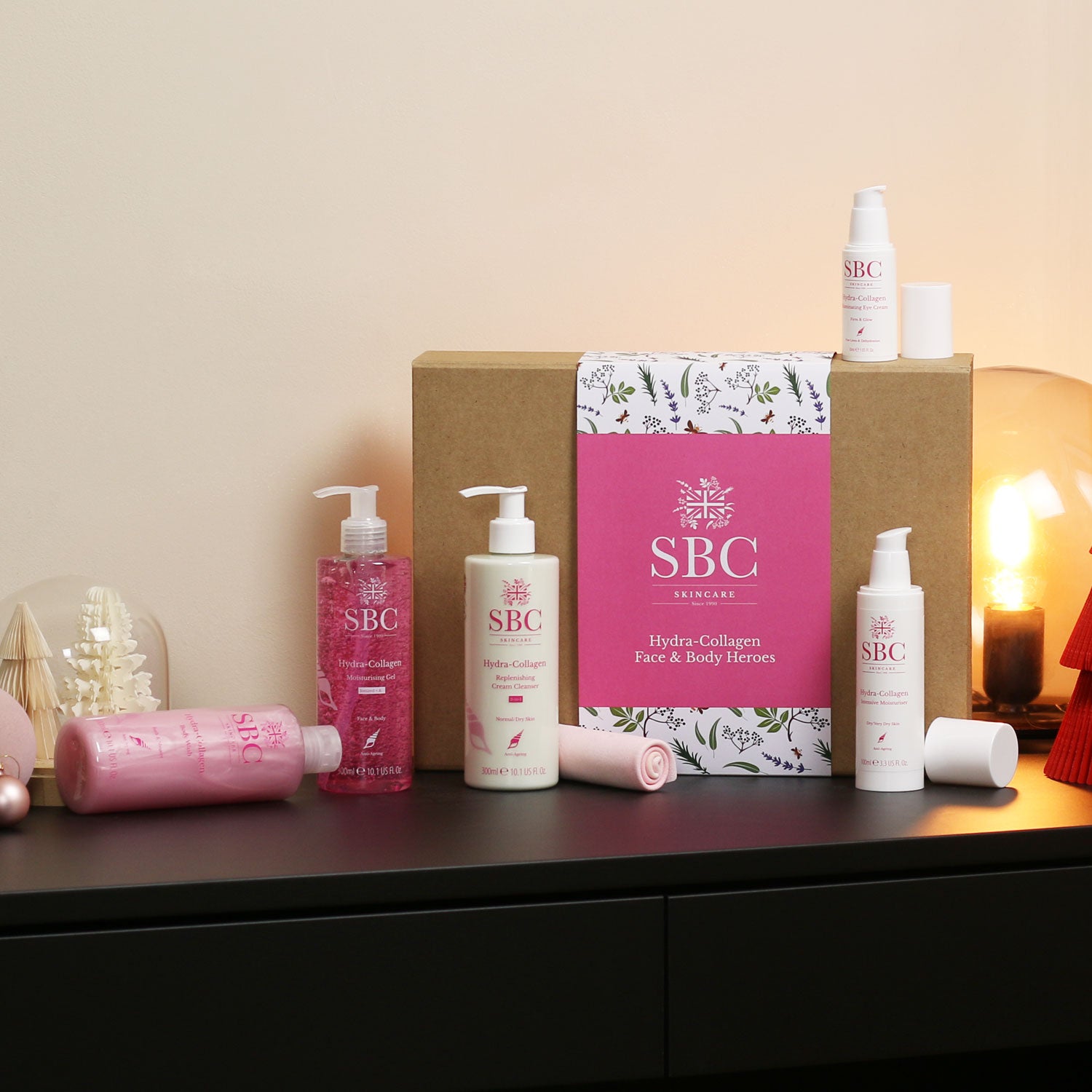 SBC Skincare’s Hydra-Collagen Face & Body Heroes gift set on a black desk with a lamp 