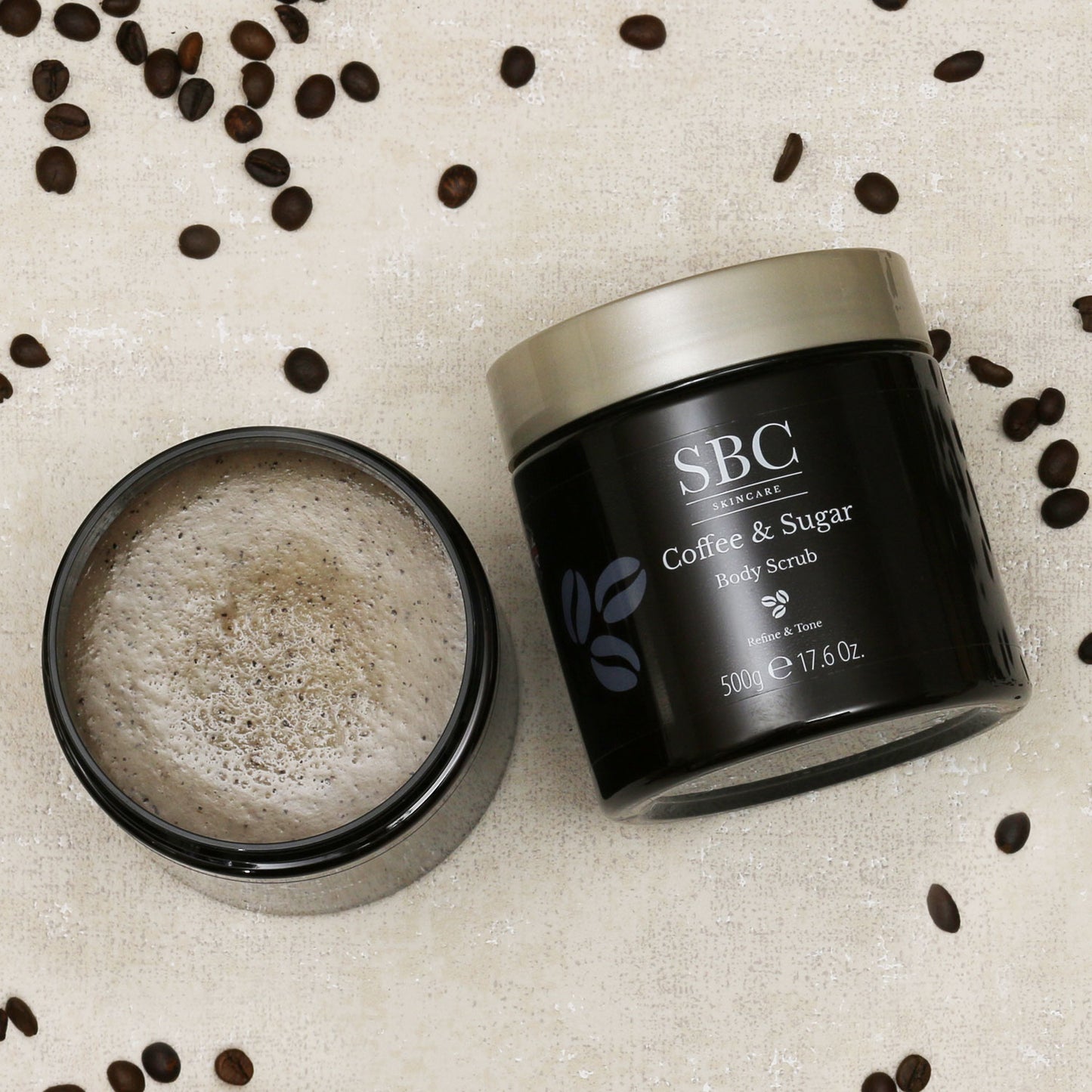 Two Coffee & Sugar Body Scrubs on a beige textured background with coffee beans sprinkled around 