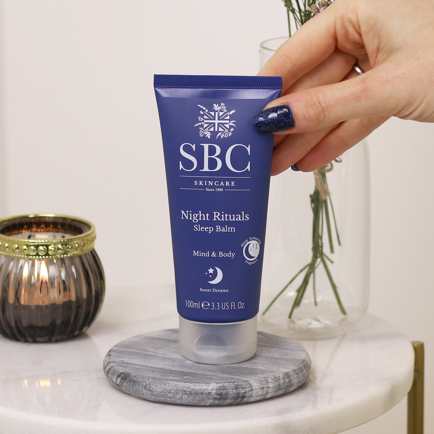 Night Rituals Sleep Balm 100ml being held by a hand with sparkly blue nails 
