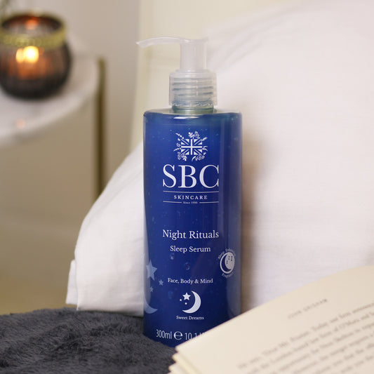 Night Rituals Sleep Serum 300ml on a bed with a book and navy throw