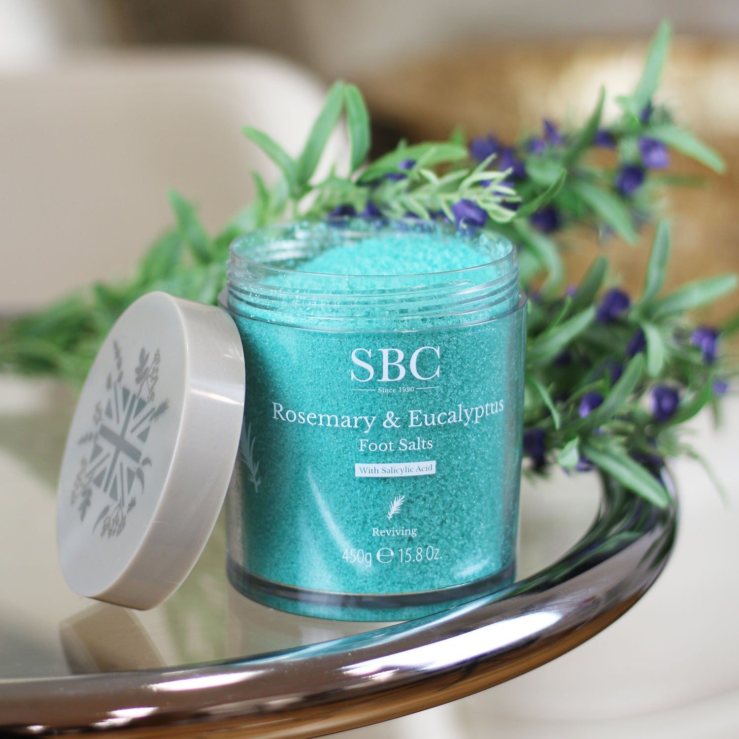 Rosemary & Eucalyptus Foot Salts on a glass table with flowering rosemary