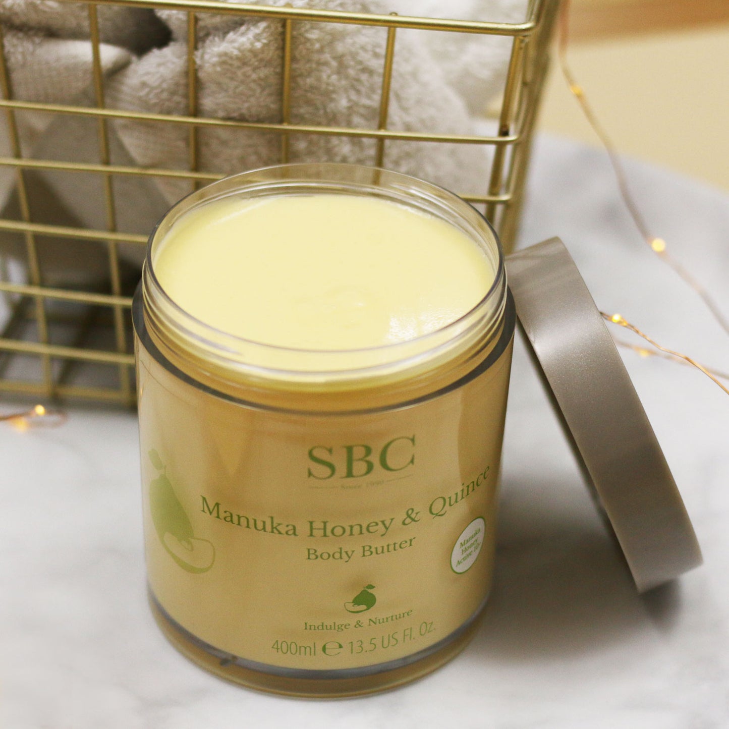 Manuka Honey & Quince Body Butter with its lid off on a marble in font of a gold basket 
